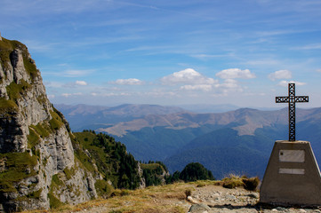 Fototapeta premium Landscape in the Carpathian Mountains with a cross marker in the foreground and a vast valley covered with trees in the background. Scene on the hiking trail of Jepii Mici in Bucegi Mountains.