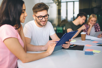 Handsome yougn man look at plastic tablet brunette hold in hands. They sit together. Another couple works behnid them. People are in white room.