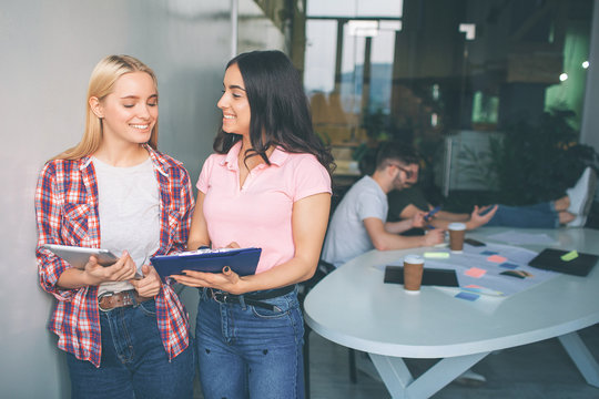 Cheerful and positive young women smile together. They stand in white room. Models hold trye and plastic tablet. Two young men sit behind them at table and work.