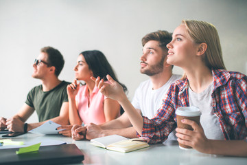 Serious and concentrated young people sit together at table. They look and think. Blonde model point with hand and hold cup of coffee.