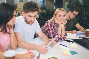 Three young peoople work seriously at table. They look down. Young blonde woman look on camera and smile. She sits between other students. All they are in same room.