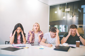Amazed and upset young blonde woman look on camera. She scream. Other people trying to concentrate. They do brainstorming. All of them sit together in white room.