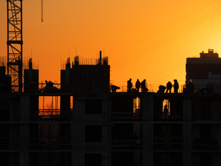 Construction of residential building. Builders go on the unfinished floor with protruding fittings. Silhouettes of workers on the background of an orange sunset.