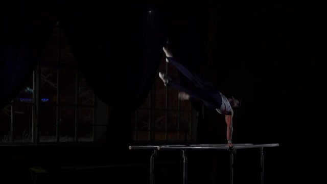 Male gymnast acrobat performs handstand on parallel bars in a dark room in slow motion sharing flips and landing on the floor