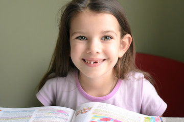 little caucasian white beautiful long haired brunette girl reading a book. smiling toothless girl with pink hair band. Smiling cute kid without front milk tooth. Kid expression portrait 