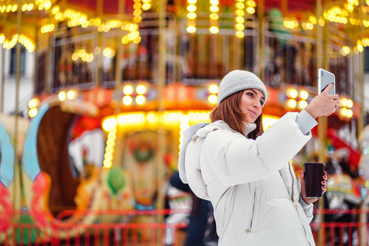 A beautiful young woman or girl in white down jacket and gray hat with coffee cup doing selfie or photographing during Christmas holidays.