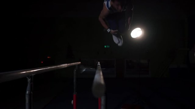 Male gymnast acrobat performs handstand on parallel bars in a dark room in slow motion sharing a somersault and landing on the floor. Training before the Olympic games. A professional athlete