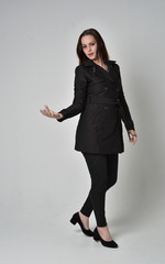 full length portrait of a brunette girl wearing long black coat, standing pose on grey studio background.