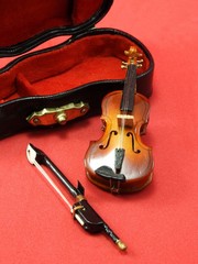 Close-up of a miniature violin with bow and black case with red lining, on a pink surface.