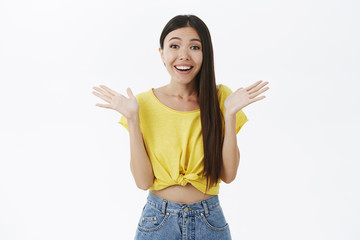 Delighted sweet asian girl with long dark hair in cropped yellow t-shirt clasping hands and smiling touched and joyful being amused and surprised with awesome big news grinning joyful over white wall