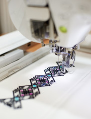 Close-up of sewing machine creating colorful abstract geometrical pattern on white fabric on blurred background. Modern technology, fashion and traditional clothing manufacturing concept.
