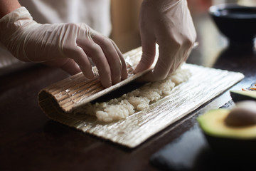 Master' s hands making a sushi roll with nori, rice, cucumber and omelet using bamboo mat. Close-up view of process. - obrazy, fototapety, plakaty