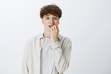 Anxious and insecure young teenage guy with curly hair in shirt biting fingernails looking unsure and nervous as frowning and looking at camera, overthinking as being concerned of things getting worse