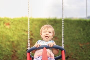 Cute happy smiling little child having fun riding on swing at children playground on sunny summer day with hig tounge out. Carefree childhood. Copy space. Close-up portrait