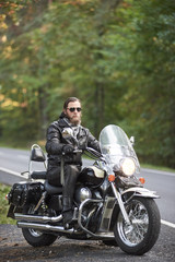 Fototapeta na wymiar Young bearded biker in black leather clothing and dark sunglasses sitting on motorcycle on country roadside on background of empty straight asphalt road and green trees bokeh foliage.