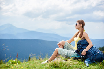 Side view of attractive woman hiker resting on grassy hill with backpack and trekking sticks, wearing sunglasses, enjoying summer cloudy day in the mountains. Outdoor activity, tourism concept