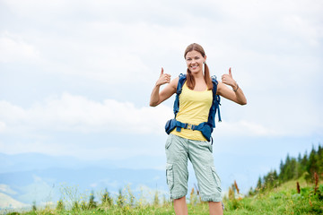 Portrait of attractive smiling woman tourist hiking mountain trail, walking on grassy hill, wearing backpack, using smiling to the camera and showing thumbs up. Outdoor activity, tourism concept