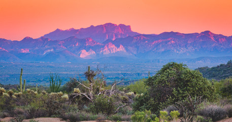 The Arizona desert mountains turn a deep reddish, orange and purple hue as the sun sets and the sky turns a soft peachy orange. Landscape photos of the desert at sunset shows a quiet and solitude 