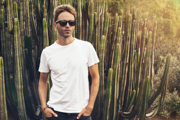 Young handsome bearded guy wearing a blank white t-shirt and sunglasses is standing in the garden...