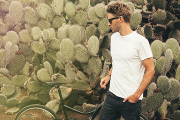 Young handsome bearded guy wearing a blank white t-shirt and sunglasses is standing in the garden background next to a big cactus plant. Horizontal mock up style © J-Project