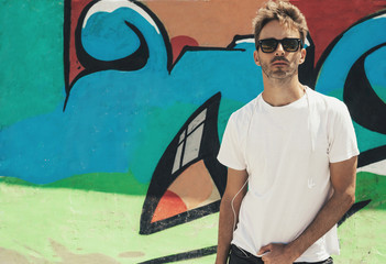 Young bearded handsome man standing next to a colorful wall background wearing an empty white t-shirt and sunglasses. Horizontal mock up style.