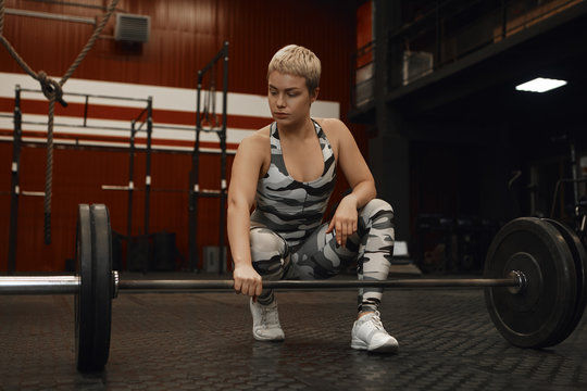 Picture of strong young woman bodybuilder working on core muscles using weights in gym. Attractive blonde female athlete with short haircut posing at fitness center with powerlifting barbell