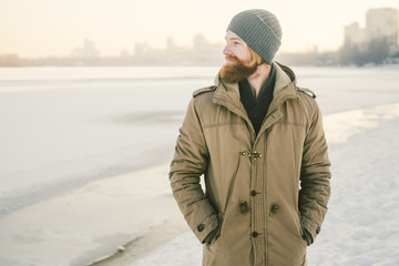 Close-up Caucasian young male red hair and beard in a hat and a park coat posing winter model against a background of a lake to freeze snow © Elizaveta