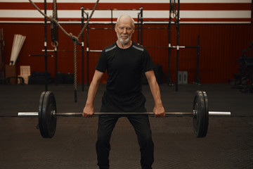 Handsome happy elderly retired male with gray beard standing in modern fitness center interior holding barbell with plates, weightlifting, enjoying physical exercise while being on retirement