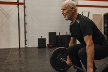 Fototapeta na wymiar Side view of confident senior male bodybuilder with gray beard doing squats indoors holding barbell. Attractive muscular retired man exercising at fitness center, shaping up, gaining srong body