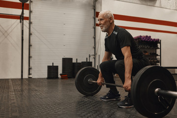 Fototapeta na wymiar Athletic unshaven grandfather wearing stylish sports clohtes and sneakers posing at modern gym with powerlifting barbell, going to do deadlift, looking ahead of him with concentrated expression
