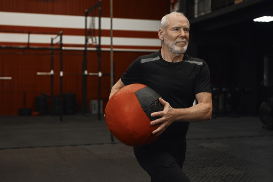 Attactive sporty senior man enjoying phycisal exercise at fitness center. Unshaven elderly retired male in sports clothes training in gym, holding weight medicine ball, doing upper body workout