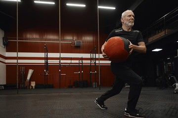 Athletic elderly male in sportswear practicing rotational lunges using medicine ball for torso and legs workout, having focused concentrated facial expression. Aging, active lifestyle and health