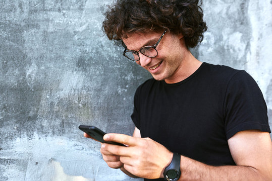 Closeup image of smiling man standing outdoors texting on mobile phone. Young male with curly hair wears spectacles resting outside in the city browsing on his cell phone on concrete gray background.