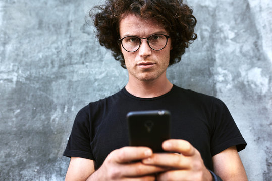 Image of handsome smart man standing outdoors texting on mobile phone. Young male with curly hair wears spectacles resting outside in the city browsing on his cell phone on concrete gray background.