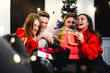 Obraz na płótnie Canvas Christmas and New Year vibes. Home decorations. Charming young woman in red sweater sits on the couch before shiny Christmas tree and opens present boxes with her friends