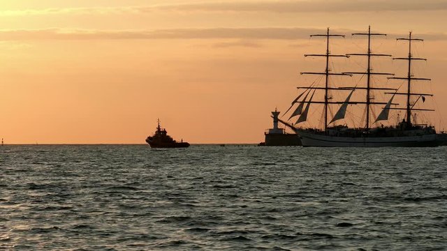 Historical tall ship replica schooner floats past the lighthouse at sea near a smaller towing boat at sunset