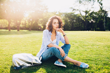 Naklejka na ściany i meble Full-length photo of cute brunette girl with short hair sitting on grass in sunlight in park . She wears white T-shirt, shirt and jeans, shoes, bag.