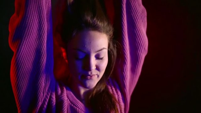 cheerful Asian teen girl dancing in pink sweater in dark under neon light red and blue
