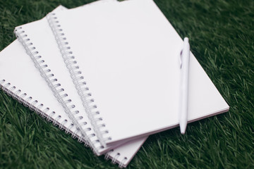 Mockup close-up of stack of white paper notebooks or notepad with white pen on the grass background