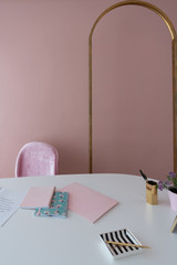Cozy pink working corner with antique gold arch frame inthe background and white working top with pink notebooks,stripe ceramic and flower in pot/ working concept / background for office space rental