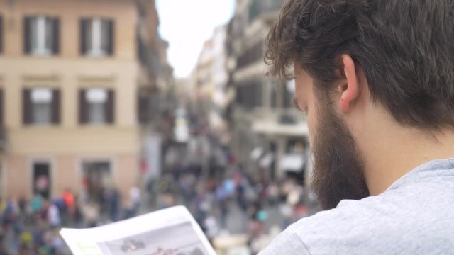 Over Shoulder View Of Man Reading Newspaper In Busy Town Square.