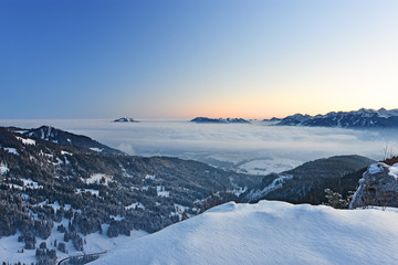 Beautiful morning mood in the Allgaeu Alps at a cold winter day. Snow-covered alpine landscape with mountains sticking out of a cloud layer and red glowing horizon before sunrise (Bavaria, Germany).