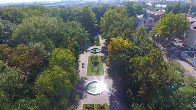 Aerial: flower beds on hill shade, people walk in Park, glare in water fountains of many green trees among in Maxim Gorky park to Krasnodar, cinematic aerial motion.