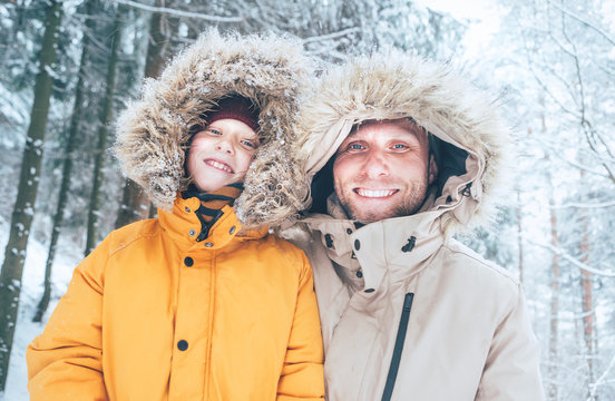 Father and son dressed in Warm Hooded Casual Parka Jacket Outerwear walking in snowy forest cheerful smiling faces portrait. Father and son relatives and winter outfit concept image.