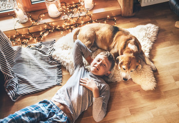 Boy lying on the floor and smiling near slipping his beagle dog on sheepskin in cozy home...