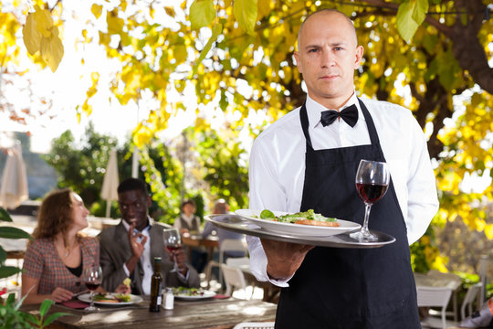 Adult waiter serves customers in a open-air restaurant
