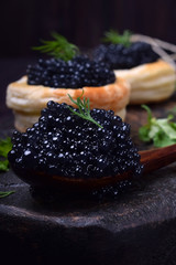 Black caviar in a wooden spoon and puff pastry tartlets on a dark board © kcuxen