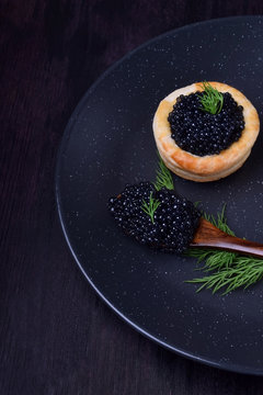 Black caviar in a wooden spoon and puff pastry tartlet on a dark plate