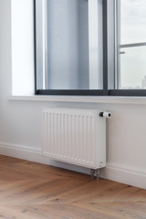 Heating white radiator with adjuster of warming in living room with large window and wooden floor.