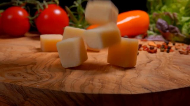 Cubes of Parmesan cheese fall to the wooden surface of the table on the background of greenery and vegetables